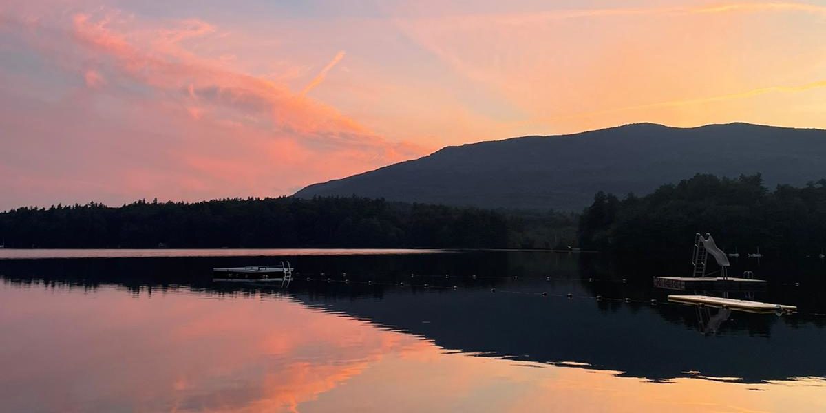 This photograph shows the view of Mt. Monadnock at sunset as seen from the swim dock. The pink and orange sunset and the mountain are reflected in the lake below.